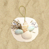 A round, beige holiday ornament featuring a shell cluster with greenery WITH A PERSONALIZED NAME printed on it.
